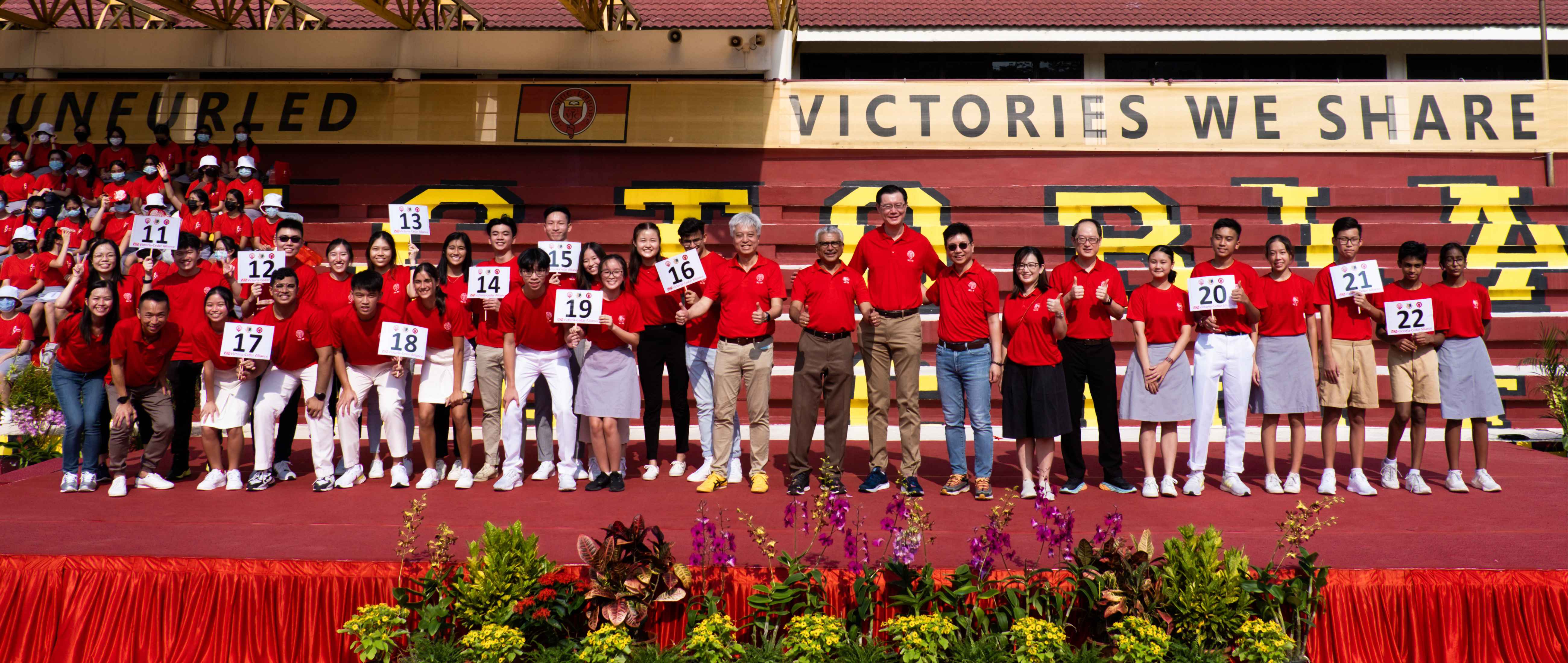 Large group in red shirts on a stadium stage holding numbered signs, with "Victories We Share" banner behind them.